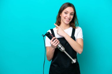 Beyaz tenli genç aşçı kadın mavi arka planda izole edilmiş el blenderi kullanıyor.