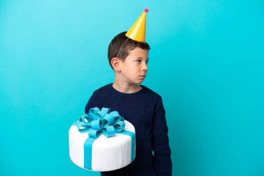 Mavi arka planda izole edilmiş doğum günü pastası tutan küçük çocuk yan tarafa bakıyor.