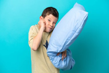 Mavi arka planda izole edilmiş pijamalı küçük çocuk hayal kırıklığına uğramış ve kulaklarını kapatmış.