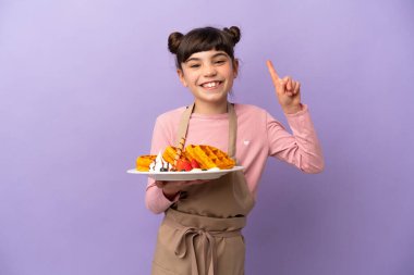 Mor arka planda izole edilmiş waffle tutan küçük beyaz kız harika bir fikre işaret ediyor.