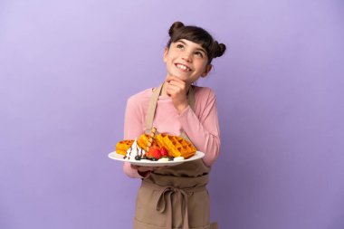 Mor arka planda izole edilmiş waffle tutan küçük beyaz kız yukarı bakıyor.