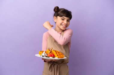 Mor arka planda izole edilmiş waffle tutan küçük beyaz kız gururlu ve kendini beğenmiş.