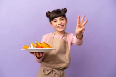 Mor arka planda gözleme tutan küçük beyaz kız mutlu ve parmaklarıyla üçe kadar sayıyor.