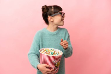 Küçük beyaz kız pembe arka planda 3 boyutlu gözlüklerle izole edilmiş ve elinde koca bir kova patlamış mısır tutuyor.