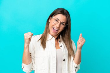 Mavi arka planda, gözlüklü ve zaferi kutlayan orta yaşlı beyaz kadın.