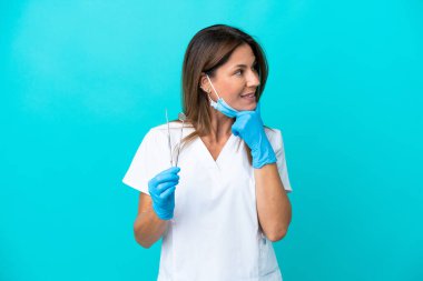 Mavi arka planda izole edilmiş orta yaşlı doktor kadın yan tarafa bakıyor ve gülümsüyor.