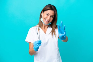 Mavi arka planda izole edilmiş orta yaşlı doktor kadın mutlu ve parmaklarıyla üçe kadar sayıyor.