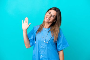 Mavi arka planda izole edilmiş orta yaşlı cerrah kadın mutlu bir ifadeyle selamlıyor.