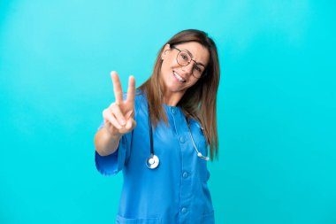 Mavi arka planda izole edilmiş orta yaşlı cerrah kadın gülümsüyor ve zafer işareti gösteriyor.