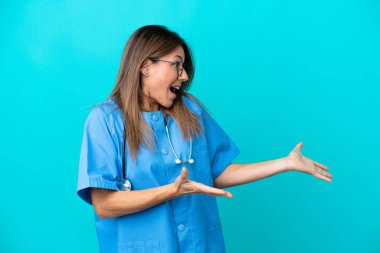 Orta yaşlı cerrah kadın, mavi arka planda, şaşırmış yüz ifadesi ile izole edilmiş.