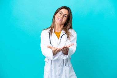 Mavi arka planda izole edilmiş orta yaşlı bir kadın doktor önlüğü giyiyor ve elinde bir şey tutuyor.