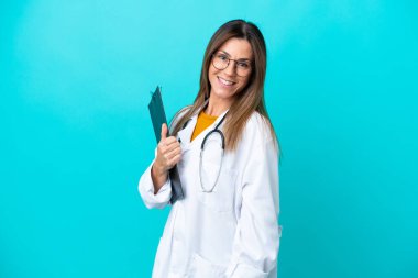 Mavi arka planda izole edilmiş orta yaşlı bir kadın doktor önlüğü giyiyor ve elinde bir klasör tutuyor.