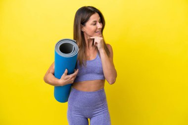 Spor kadını yoga derslerine giderken elinde bir minder, sarı olmayan bir arka plan, yana bakıyor ve gülümsüyor.