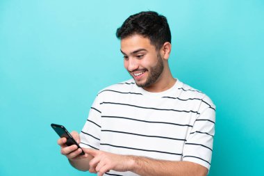 Genç Brezilyalı adam mavi arkaplanda izole edilmiş cep telefonuyla mesaj ya da e-posta gönderiyor