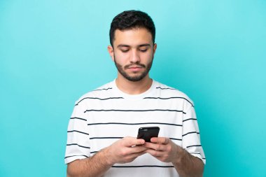 Genç Brezilyalı adam mavi arka planda cep telefonu kullanarak izole edilmiş.