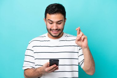 Genç Brezilyalı adam mavi arka planda izole edilmiş ve parmakları çapraz cep telefonu kullanıyor.