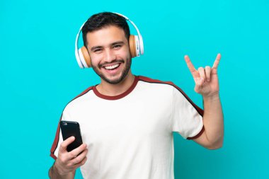 Genç Brezilyalı bir adam mavi arka planda izole edilmiş, hareketli bir rock hareketi yaparak müzik dinliyor.