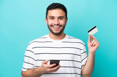 Genç Brezilyalı bir adam mavi arka planda izole edilmiş cep telefonuyla alışveriş yapıyor ve elinde şaşırmış bir ifadeyle bir kredi kartı tutuyor.