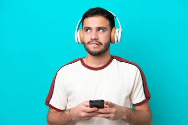 Genç Brezilyalı bir adam mavi arka planda izole edilmiş. Cep telefonuyla müzik dinliyor ve düşünüyor.