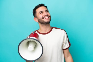 Genç Brezilyalı bir adam mavi arka planda izole edilmiş elinde bir megafonla yukarı bakıyor ve gülümsüyor.