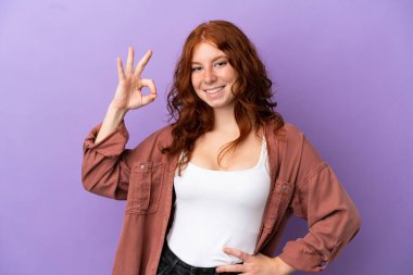 İzole edilmiş mor arka planda genç bir kızıl kız parmaklarıyla tamam işareti gösteriyor.