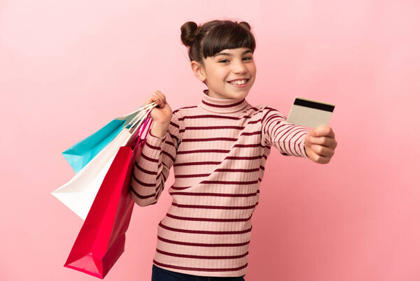 Маленькая белая девочка, изолированная на розовом фоне, держащая сумки с покупками и кредитку