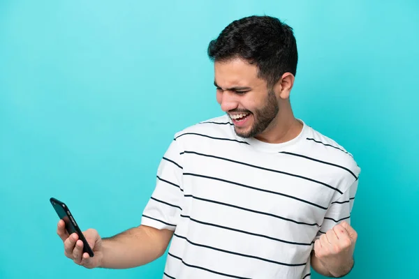 Junger Brasilianer Isoliert Auf Blauem Hintergrund Mit Handy Und Siegesgeste — Stockfoto