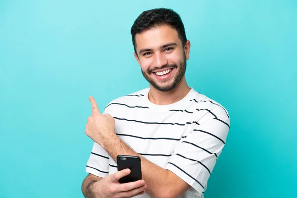 Νεαρός Βραζιλιάνος Απομονωμένος Μπλε Φόντο Χρησιμοποιώντας Κινητό Τηλέφωνο Και Δείχνοντας — Φωτογραφία Αρχείου
