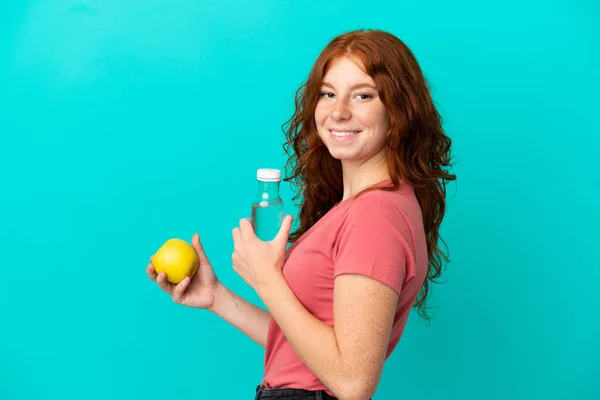 十代の赤毛の女の子はリンゴと水のボトルで青の背景に隔離されました — ストック写真