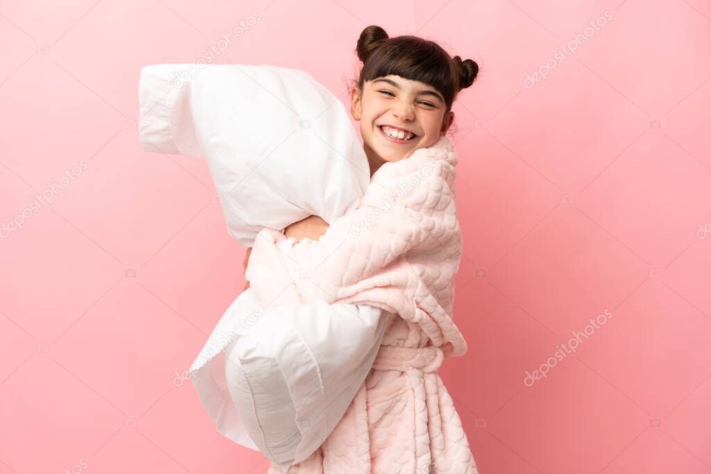 Little caucasian girl isolated on pink background in pajamas and holding a pillow with happy expression