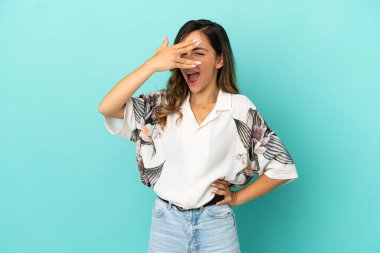 İzole edilmiş mavi arka planda genç bir kadın. Gözleri elleriyle kaplıyor ve gülümsüyor.