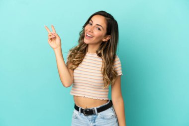 İzole edilmiş mavi arka planda genç bir kadın gülümsüyor ve zafer işareti gösteriyor