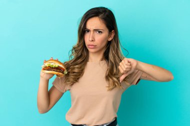 Soyutlanmış arka planda hamburger tutan genç bir kadın, olumsuz bir ifadeyle baş parmağını aşağı gösteriyor.