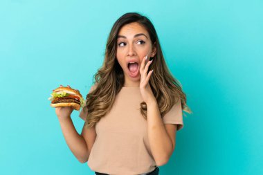 Elinde hamburger tutan genç bir kadın şaşırmış ve şok olmuş bir yüz ifadesiyle izole edilmiş bir arka plan.