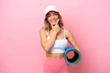 Genç sporcu kadın, elinde minderle yoga derslerine gidip gülümsüyor.