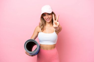 Genç sporcu kadın yoga derslerine giderken elinde bir minder, gülümsüyor ve zafer işareti gösteriyordu.