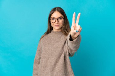 Mavi arka planda izole edilmiş genç Litvanyalı kadın gülümsüyor ve zafer işareti gösteriyor.