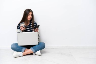 Young caucasian woman with a laptop sitting on the floor isolated on white background holding coffee to take away and a mobile