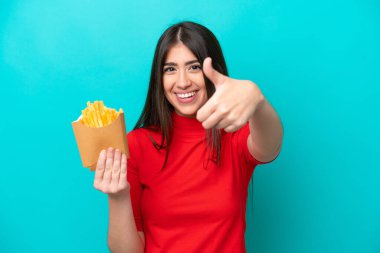 Genç beyaz kadın patates kızartması yakalıyor. Mavi arka planda izole edilmiş. Başparmakları havada çünkü iyi bir şey oldu.