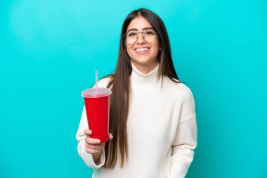 Mavi arka planda izole edilmiş gazoz içen genç beyaz kadın kolları kalçasında ve gülümsüyor.