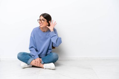 Beyaz arka planda izole bir şekilde yerde oturan beyaz bir kadın elini kulağına koyarak bir şeyler dinliyor.