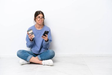 Beyaz arka planda izole bir şekilde yerde oturan beyaz tenli genç bir kadın elinde kahve ve bir şeyler düşünürken bir cep telefonu tutuyordu.