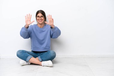 Beyaz arka planda yerde oturan beyaz tenli genç kadın parmaklarıyla dokuz kişi sayıyor.