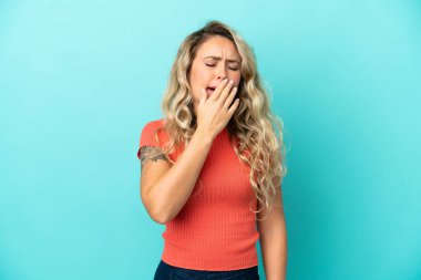 Genç Brezilyalı bir kadın mavi arka planda izole edilmiş esniyor ve ağzını eliyle açık tutuyor.