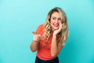 Mavi arka planda izole edilmiş genç Brezilyalı kadın bir ürünü sunmak ve bir şeyler fısıldamak için kenarı işaret ediyor.