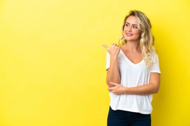 Sarı arka planda izole edilmiş genç Brezilyalı kadın bir ürün sunmak için kenarı işaret ediyor.