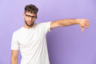 Mor arka planda izole edilmiş beyaz bir adam negatif ifadeyle baş parmağını aşağı gösteriyor.