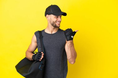 Young sport blonde man with sport bag isolated on yellow background pointing to the side to present a product