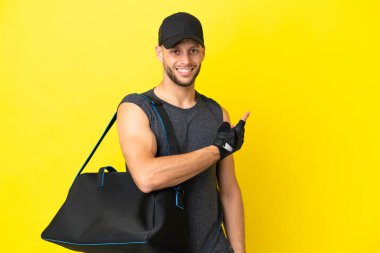 Young sport blonde man with sport bag isolated on yellow background pointing back