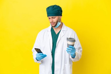 Sarı arka planda izole edilmiş yeşil üniformalı cerrah. Elinde kahve ve cep telefonu var.